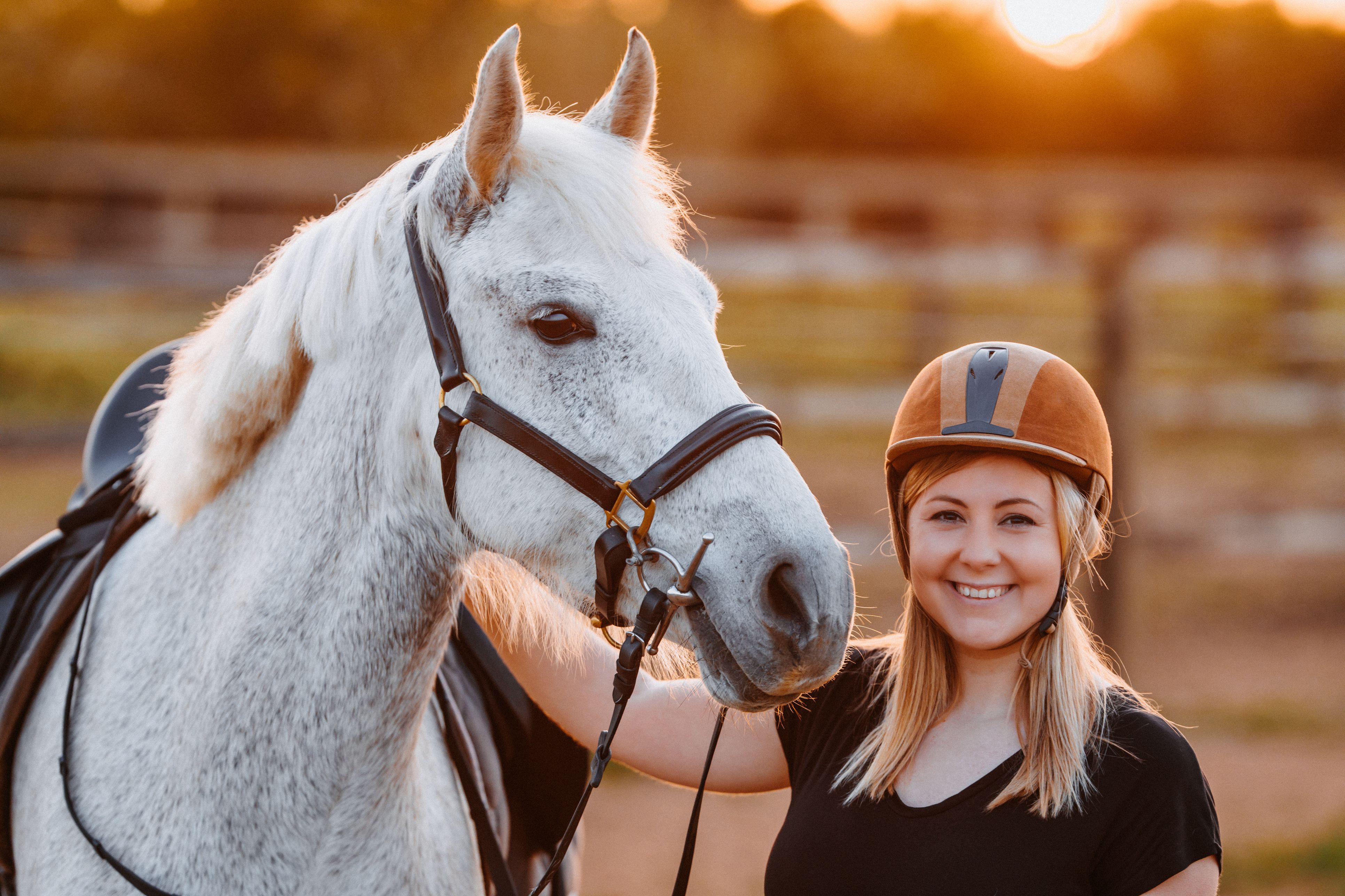 Eine blonde Frau mit Reiterhelm steht lächelnd neben einem weißen Pferd auf der Koppel.
